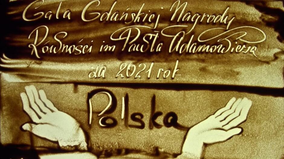 Obraz z piachu w różnych odcieniach - od jasnego żółtego po brązowy. Górna część ma ozdobny napis Gala Gdańskiej Nagrody Równości im. Pawła Adamowicza za rok 2021. W dolnej części znajduje się wizerunek pary rozłożonych i skierowanych w górę ludzkich dłoni. Między nimi znajduje się napis Polska