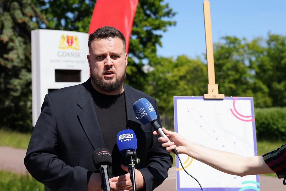 Na zdjęciu mężczyzna w czarnej marynarce wypowiada się do mediów podczas konferencji prasowej, przed kilkoma mikrofonami. W tle widać tablicę z mapą oraz oznaczenia miasta Gdańsk, co sugeruje, że wydarzenie dotyczy spraw lokalnych lub infrastrukturalnych.