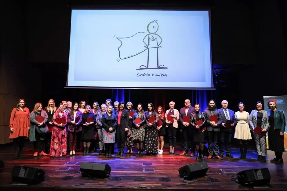 Na zdjęciu widzimy grupę ludzi stojących na scenie podczas uroczystej ceremonii, trzymających czerwone teczki z dokumentami. W tle widoczny jest ekran z rysunkiem postaci w pelerynie i napisem Ludzie z misją, co sugeruje, że wydarzenie jest poświęcone wyróżnieniu osób za ich wyjątkowe osiągnięcia lub zaangażowanie w ważne projekty.